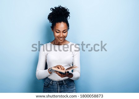Attractive, young woman with a smile reads an interesting book on a background of blue wall