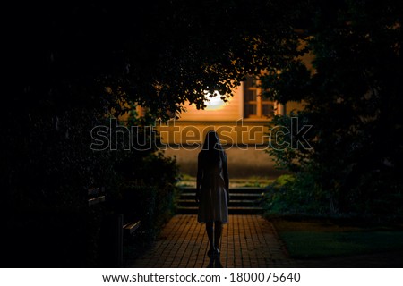 Young alone woman in dress walking on sidewalk through dark park to home in summer black night. Scary moment and gloomy atmosphere. Back view. Royalty-Free Stock Photo #1800075640