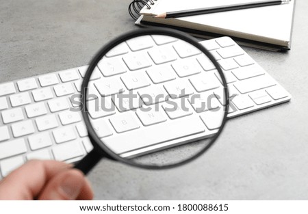 Woman holding magnifier glass near keyboard at light grey stone table, closeup. Find keywords concept