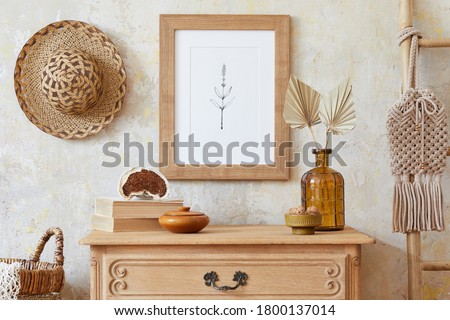 Stylish boho interior of living room with brown mock up poster frame, elegant accessories, flowers in vase, wooden shelf and hanging rattan hut. Minimalistic concept of home decor. Template. 