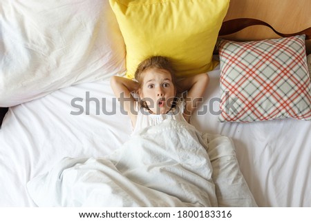 A little girl with fair hair lies on a large bed. Laugh, watch cartoons on your phone and tablet. Play on your gadget. White linens. Children are Europeans.