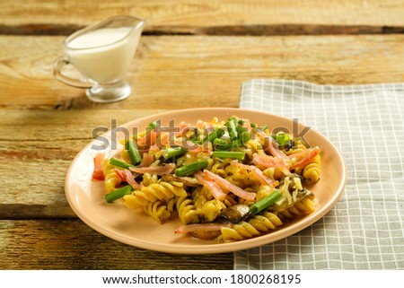 a plate with pasta with ham and mushrooms in a creamy sauce on the table with a fork on a napkin next to the cream.