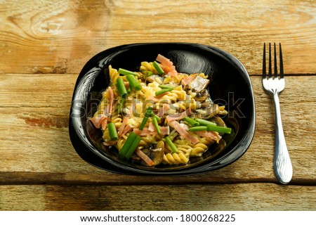 Black plate with pasta with ham and mushrooms in a creamy sauce on the table with a fork.