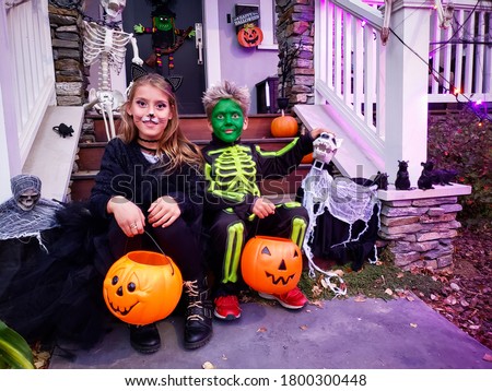 Halloween kids with orange Jack o Lantern buckets, girl with black cat costume and boy with green zombie, skeleton costume in front of Halloween decorated house are celebrating Halloween evening night