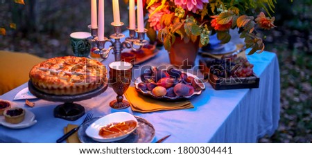 Autumn evening photo shoot - romantic dinner outdoors. Table with tablecloth and decoration - pie, figs, glasses, plates, table setting and candelabra with candles. Fall flowers dahlia bouquet.