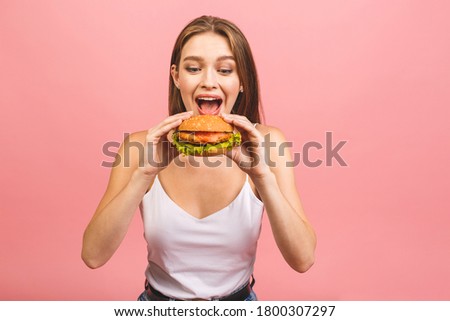 Portrait of young beautiful hungry woman eating burger. Isolated portrait of student with fast food over pink background. Diet concept. 