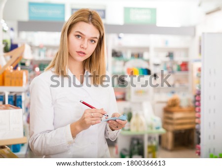 Diligent friendly smiling female pharmacist noting assortment of drugs in pharmacy. High quality photo