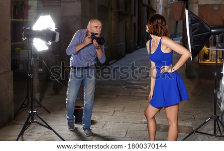 Photographer using professional camera and light equipment for taking pictures of young woman in blue dress on town street