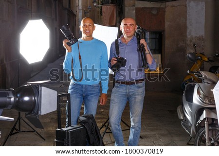 Portrait of two positive photographers standing with cameras among professional photo equipment on old city street