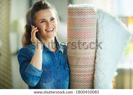 happy modern housewife in jeans shirt and white pants with white carpet talking on a smartphone in the modern house in sunny day.