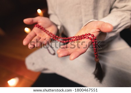 close-up man in clothes for practice and meditation sits in a lotus pose and holds red rosary to concentrate attention in a wooden room with dim light