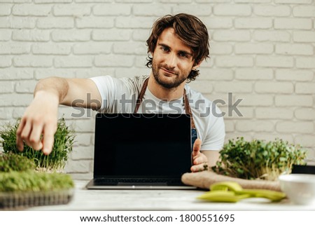 Man farmer shows black screen of laptop and sits at the table with sprouts