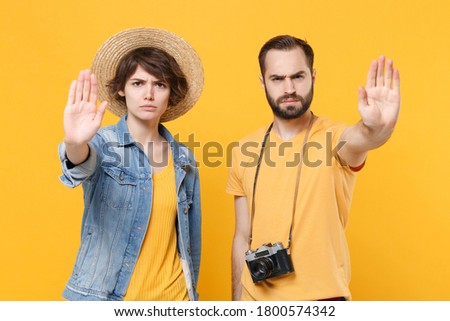Serious tourists couple friends guy girl in summer clothes with photo camera isolated on yellow background. Passenger traveling abroad on weekends. Air flight journey. Showing stop gesture with palms