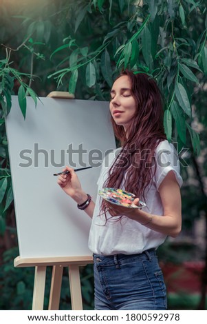 Woman artist, summer in park, background easel brush and color palette with paints. Eyes closed, dreams and fantasies. Emotions of tenderness and pleasure. Green leaves of trees.