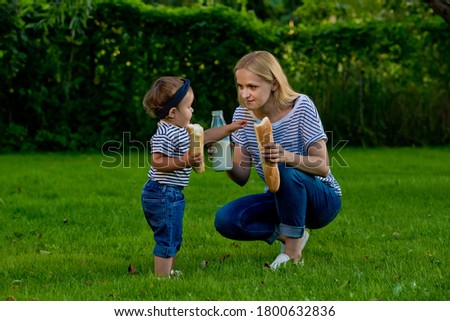A young woman in jeans and a striped T-shirt gives her daughter milk from a glass bottle. Family picnic.