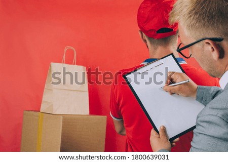 A man with a cardboard box and documents in his hands, hands over the parcel to the client. Delivery service concept on red background