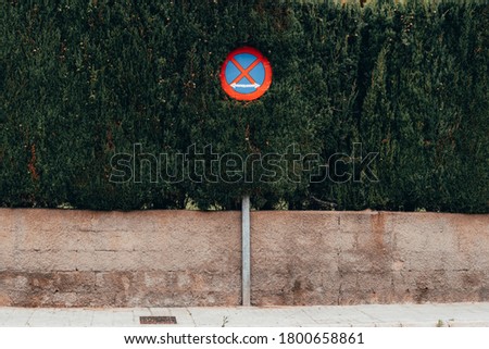 Round traffic sign with a red cross embedded in a lush bush on the sidewalk, the sign means a parking ban