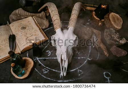 White goat scull with horns, open old book (text - untranslatable, fictional language), black candles on witch table. Occult, esoteric, divination and wicca concept. Halloween, Day of the dead concept