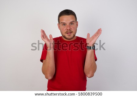 Surprised terrified Young caucasian businessman  wearing red shirt over white background Gestures with uncertainty, stares at camera, puzzled as doesn't know answer on tricky question, People, 