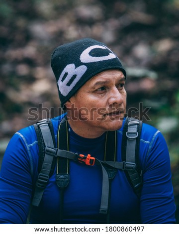 Old white man looking to the horizon on a green and cold forest background. Adult male with black bennie, black eyes and hiking gear. 