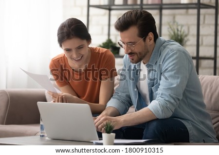 Happy millennial Caucasian couple sit on couch at home look at laptop screen shopping online together, smiling man and woman browse internet pay bills on internet on computer, use modern gadget Royalty-Free Stock Photo #1800910315