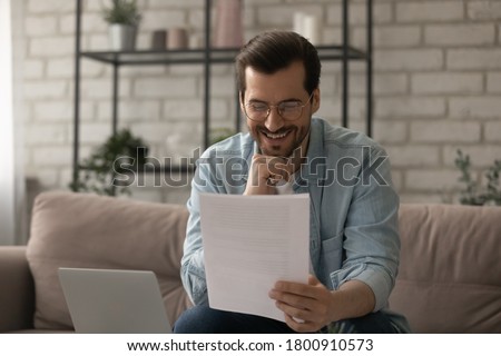 Happy young Caucasian man sit on couch at home read good news in postal paper letter, smiling millennial male in glasses get pleasant message or promotion offer in post paperwork correspondence Royalty-Free Stock Photo #1800910573