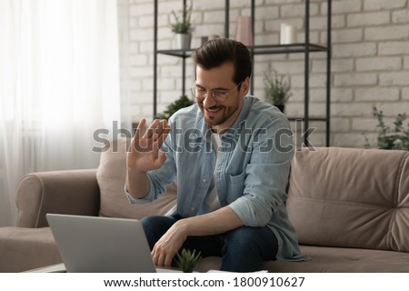 Smiling young Caucasian man in glasses sit on couch at home wave greeting talking on video call on laptop, happy male have webcam virtual conference or interview on computer, digital concept Royalty-Free Stock Photo #1800910627