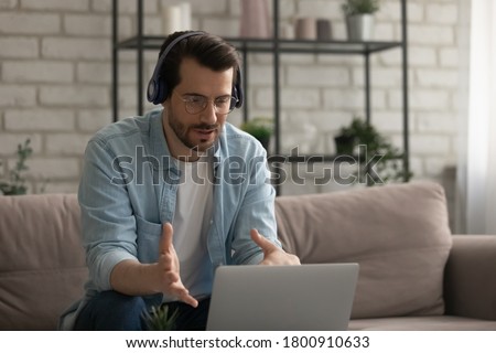 Young Caucasian man in headphones glasses sit on sofa at home talk on video call on laptop, millennial male in earphones have webcam virtual online conference conversation on computer with client Royalty-Free Stock Photo #1800910633
