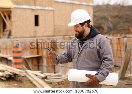 Caucasian builder using mobile phone internet and holding drawing plan at construction site, wearing hardhat. Concept of architectural profession and house building. Royalty-Free Stock Photo #1800982678