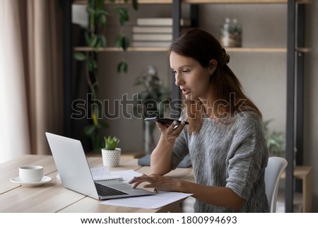 Focused young Caucasian woman sit at desk at home work on laptop record audio message on cellphone, millennial female use computer browsing, activate virtual digital voice assistant on smartphone