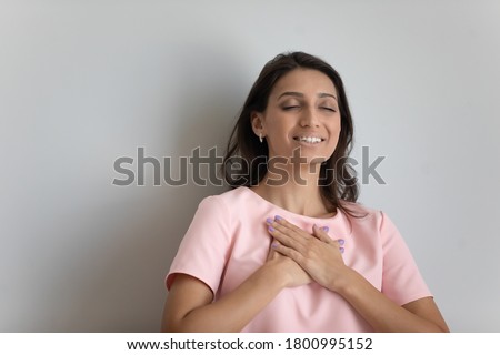 Grateful Arabic indian young woman isolated on grey studio background hold keep hands at heart chest feel thankful, happy ethnic millennial girl show appreciation gratitude, believer, faith concept