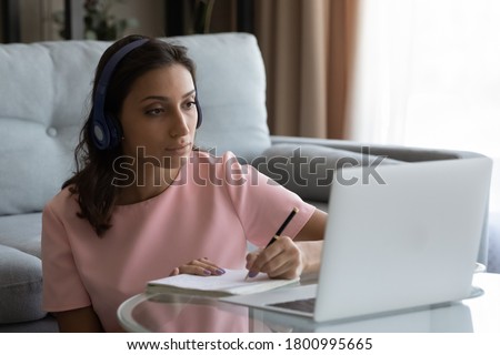 Concentrated young Arabic indian woman in headphones look at laptop screen make notes watching webinar at home, focused arab female in earphones study online on computer, distant education concept Royalty-Free Stock Photo #1800995665