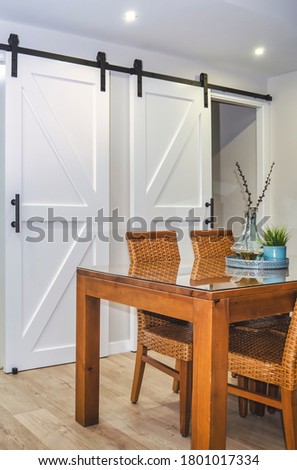 Selective focus on a bright dining room with a wooden table and rattan chairs. White sliding barn doors in the background. Royalty-Free Stock Photo #1801017334