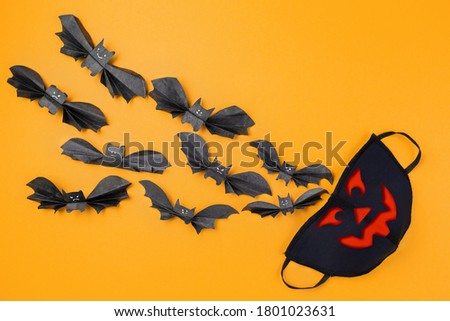 Reusable anti-virus mask with print of jack lantern surrounded by paper bats. Orange background. Copy space. Flat lay. The concept of protection from the virus during Halloween