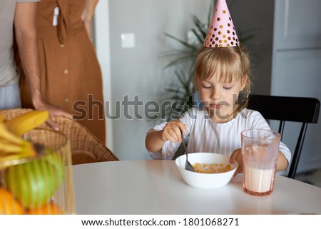 pretty adorable girl sitting in the kitchen alone, in party hat, before birthday party at home. eat, have meal in the morning