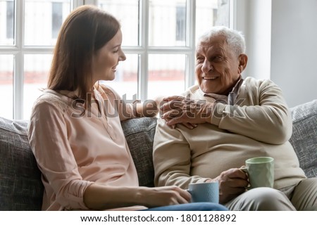Smiling young woman sitting on sofa with happy older retired 70s father, enjoying pleasant conversation with cup of coffee tea together in living room, mature parents and grown children communication. Royalty-Free Stock Photo #1801128892