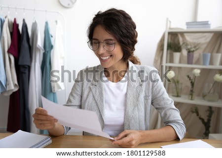 Happy young beautiful female clothing designer beginner holding paper letter, reading invitation for fashion show in modern showroom. Smiling brand owner businesswoman excited by good news in office.