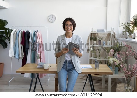 Portrait of young decorator in glasses leaning at table, holding computer tablet in modern atelier. Smiling professional florist businesswoman entrepreneur looking at camera, posing in showroom. Royalty-Free Stock Photo #1801129594