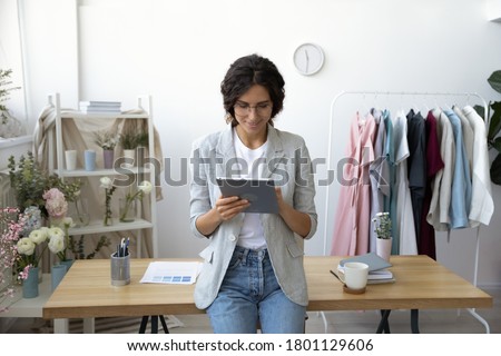 Smiling young attractive professional decorator leaning at table, communicating with clients on computer tablet indoors or using applications for creating sketching new clothing collection in office.