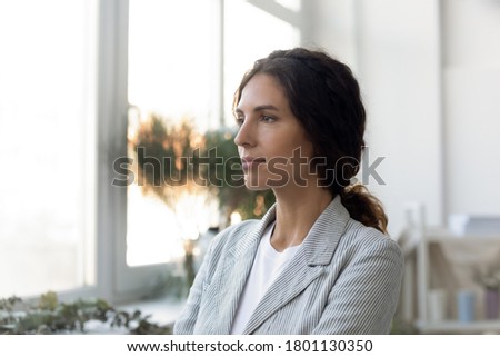 Head shot side view young thoughtful businesswoman standing near window, looking away in distance. Lost in thoughts millennial professional decorator thinking of business problems or brand development