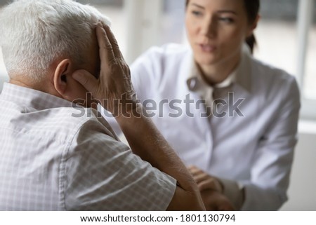 Stressed older senior patient touching head, complaining about migraine illness, meeting doc nurse at home. Professional female general practitioner treating unhealthy elderly mature retired man. Royalty-Free Stock Photo #1801130794