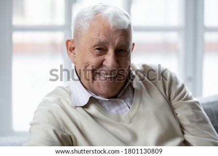 Head shot portrait of happy elderly 80s retired man relaxing on couch indoors. Smiling old mature grandfather looking at camera, feeling healthy alone at retirement house, good mood healthcare concept