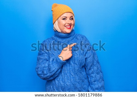 Young beautiful blonde plus size woman wearing blue casual turtleneck sweater and wool hat smiling cheerful pointing with hand and finger up to the side