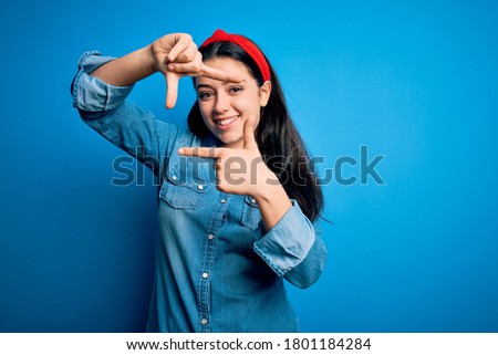 Young brunette woman wearing casual denim shirt over blue isolated background smiling making frame with hands and fingers with happy face. Creativity and photography concept.