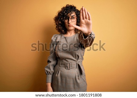 Beautiful arab business woman wearing dress and glasses standing over yellow background doing stop sing with palm of the hand. Warning expression with negative and serious gesture on the face.