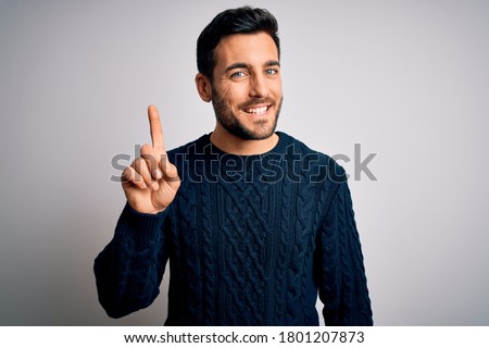 Young handsome man with beard wearing casual sweater standing over white background showing and pointing up with finger number one while smiling confident and happy. Royalty-Free Stock Photo #1801207873