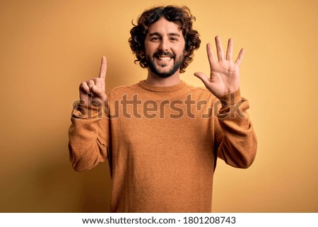 Young handsome man with beard wearing casual sweater standing over yellow background showing and pointing up with fingers number six while smiling confident and happy.