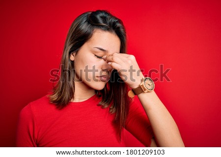 Young beautiful brunette girl wearing casual t-shirt over isolated red background tired rubbing nose and eyes feeling fatigue and headache. Stress and frustration concept.