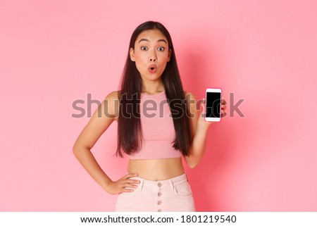 Technology, communication and online lifestyle concept. Intrigued and curious asian girl looking wondered while asking question about mobile phone app, pointing at smartphone screen, pink background