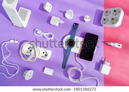 The concept of wireless charging of mobile gadgets. The smartphone and smartwatch are on the wireless charging pad. There are various adapters, cables and chargers nearby. Colored paper background. Royalty-Free Stock Photo #1801286272